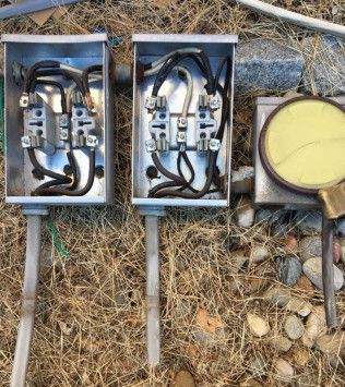 Metal electrical boxes with wires and a meter in an outdoor setting on dry grass and rocks.