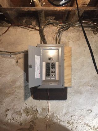 Electrical panel with open door mounted on a concrete wall in a basement. Wires are visible.