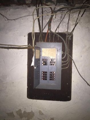 Electrical panel with exposed wires on a weathered basement wall.