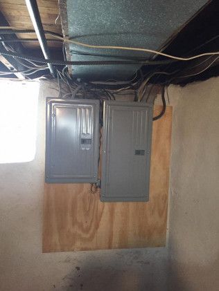 Two gray electrical panels mounted on plywood in a basement. Wires and pipes are visible above.