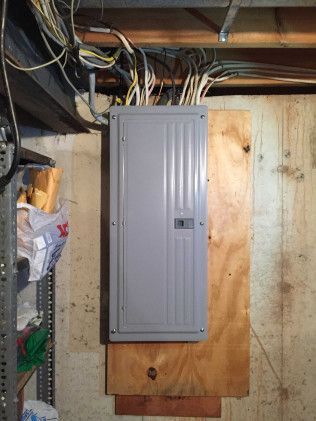 Electrical panel mounted on wood in a basement with exposed wiring.