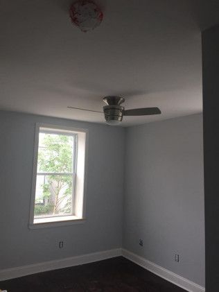 A room with a window, ceiling fan, and smoke detector. Light blue walls and dark wood floor.