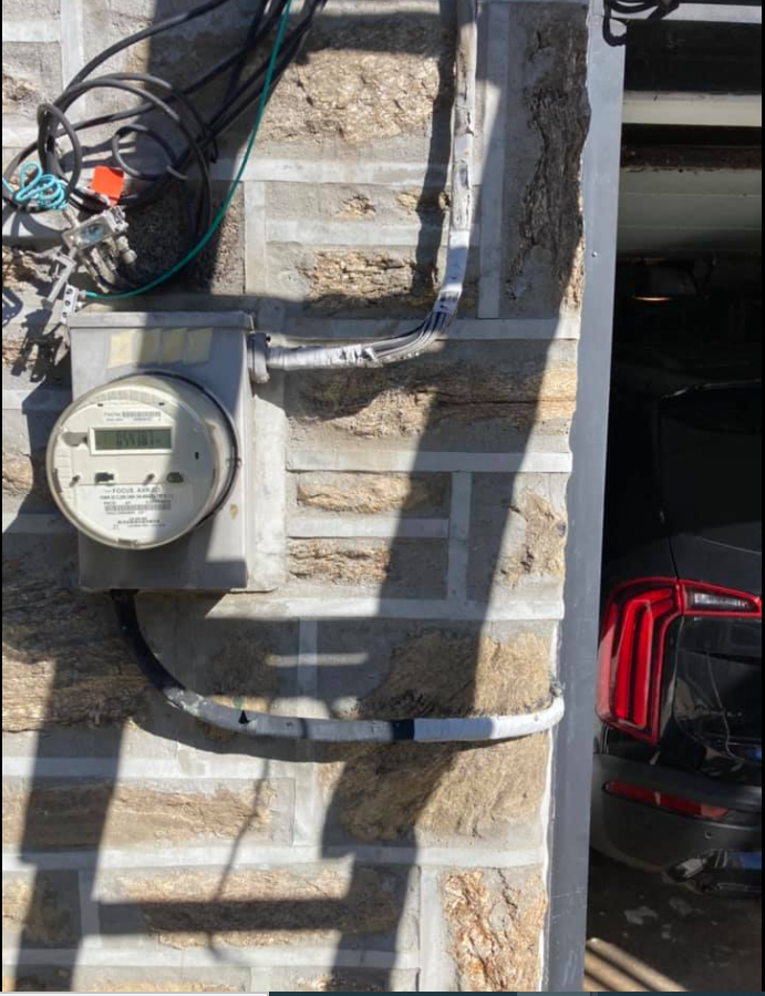 Electric meter on stone wall near a garage. Wires and conduit are visible.
