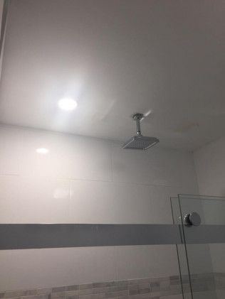 Shower with square showerhead and overhead lighting. White tile walls with a glass door.
