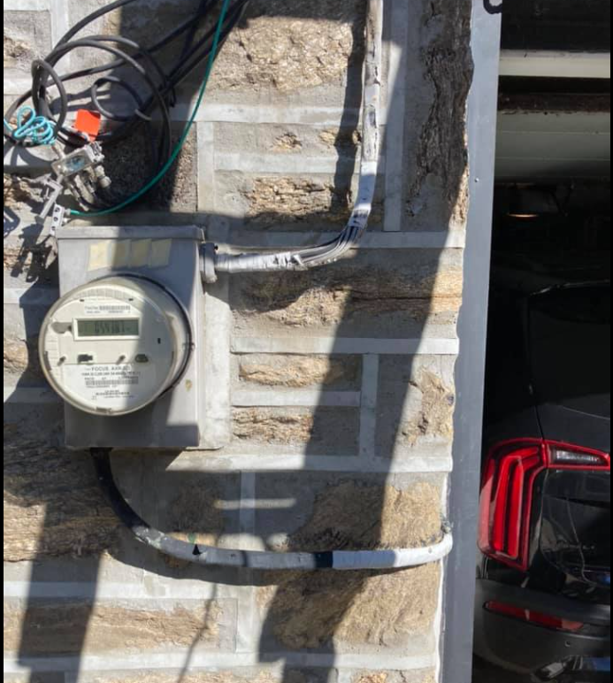 Electric meter and wiring on a beige brick wall next to a garage entrance with a red car tail light visible.