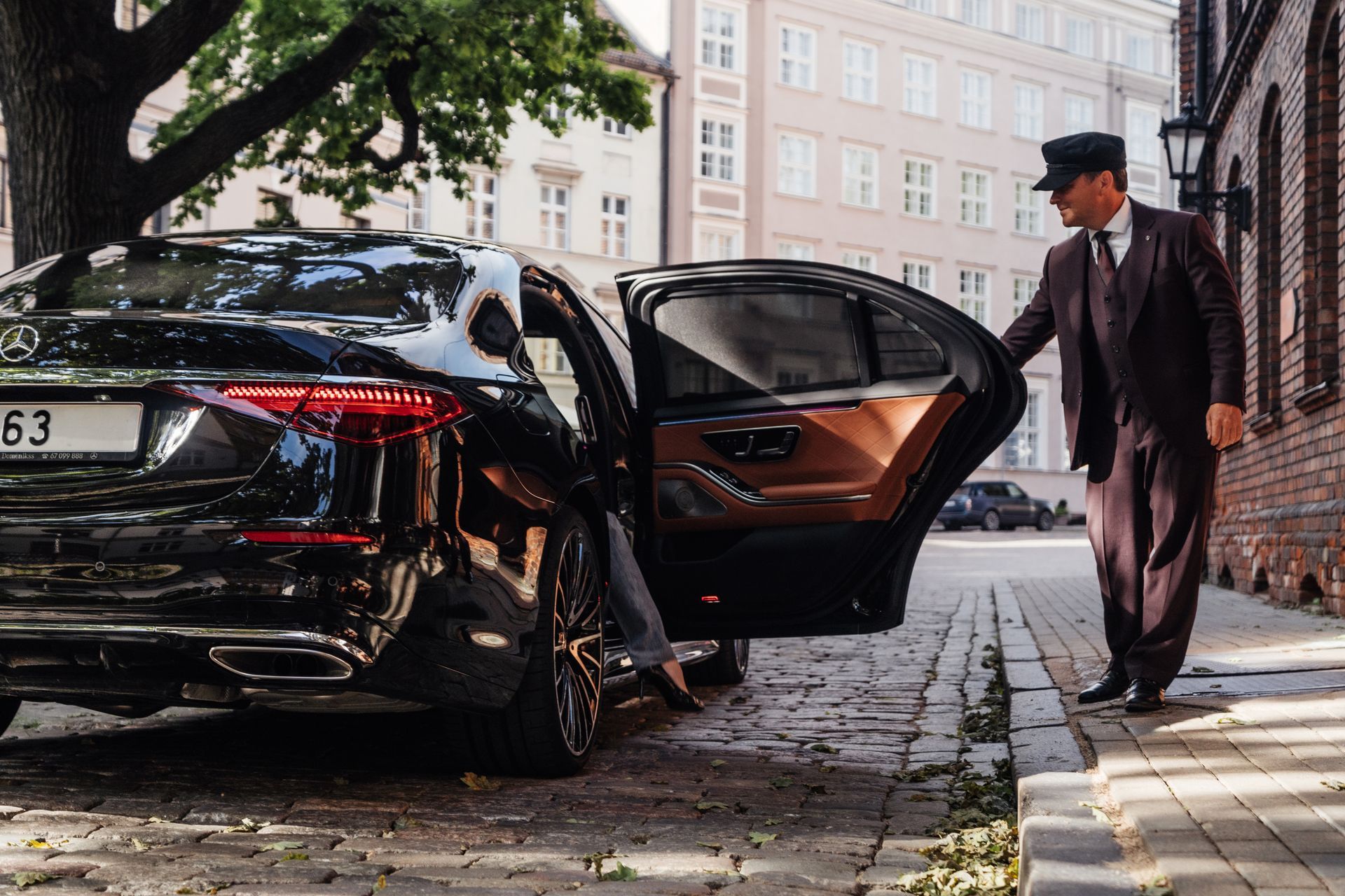 Chauffeur in a dark suit opens the car door of a black luxury sedan.