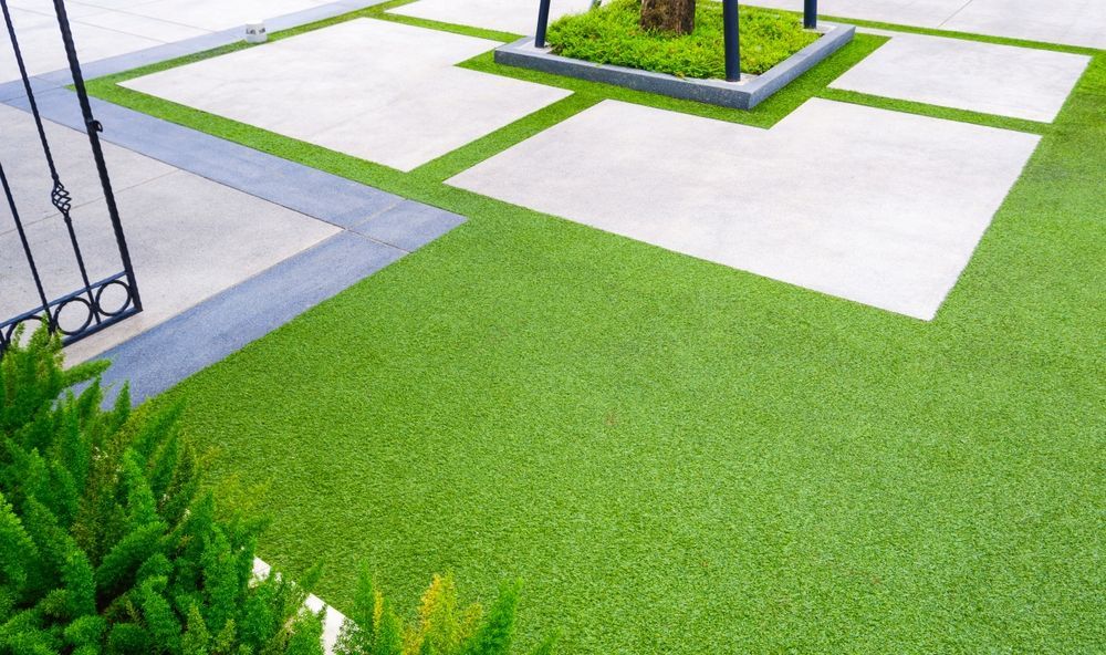 Green artificial turf contrasts with concrete slabs in an outdoor space, tree in center.