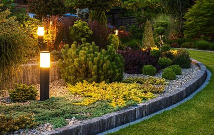 Illuminated garden bed with a variety of shrubs and ornamental grasses, edged with stone and illuminated by vertical lights.