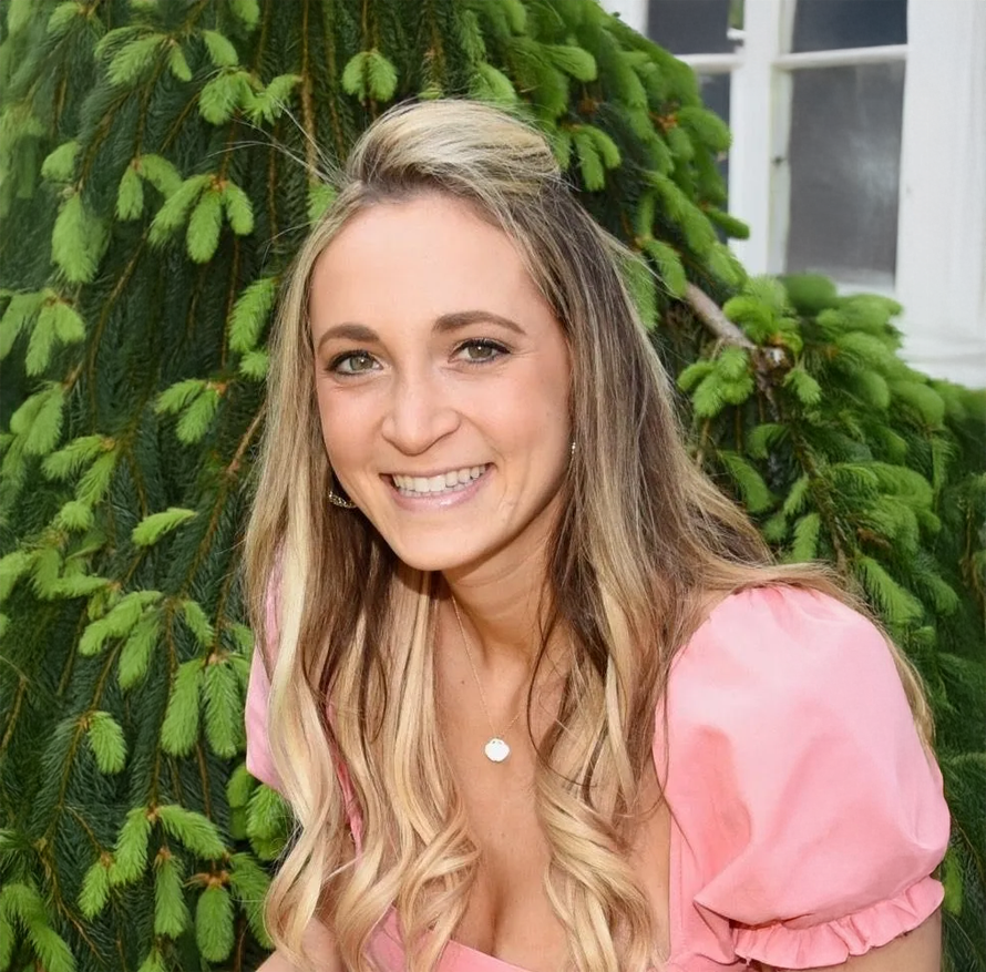 A woman in a pink top is smiling in front of a tree.