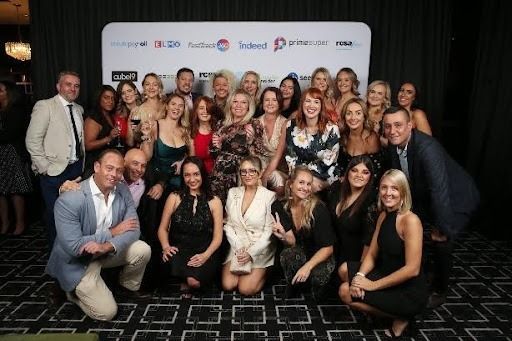Group of people at an event, smiling. Formal attire, dark background, company logos visible.