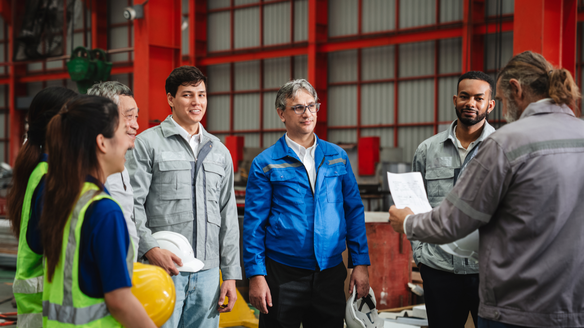 Group of people in work attire, listening to a person holding papers in a factory.