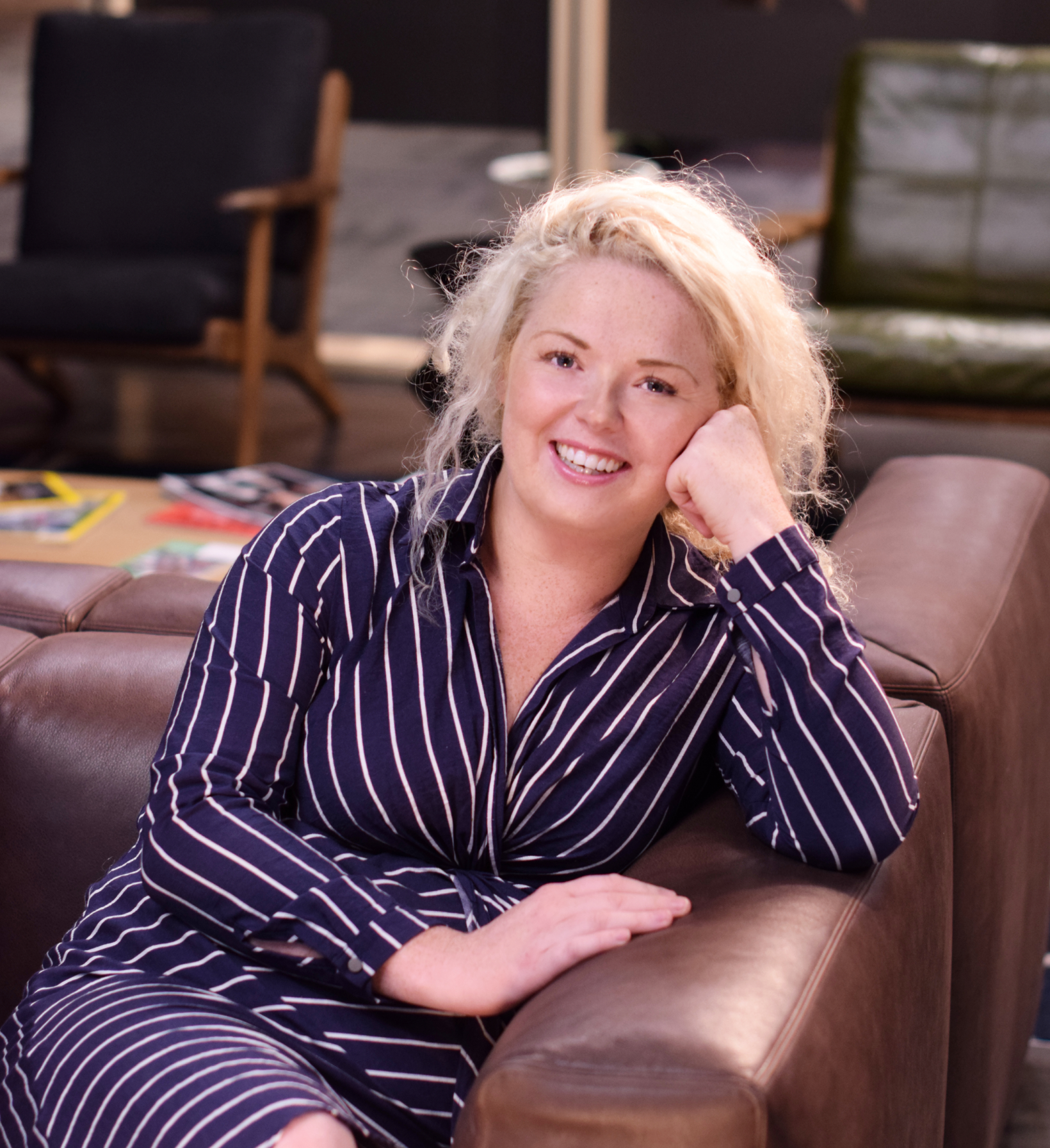 Woman with blonde hair wearing a striped shirt dress, smiling, sitting on a leather sofa.