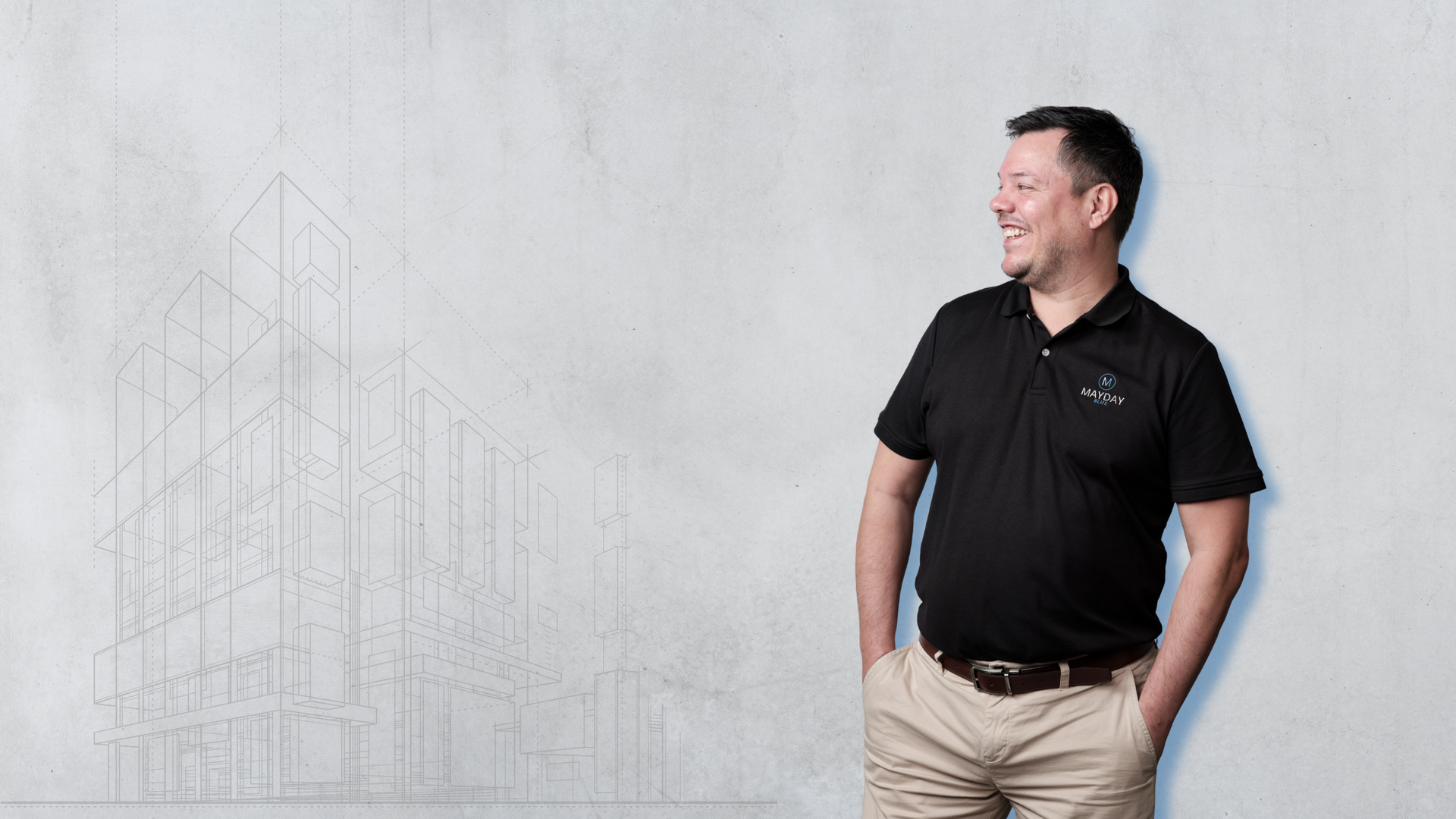 Man in black polo shirt and khakis smiling, hands in pockets, looking right, with building sketch on wall.