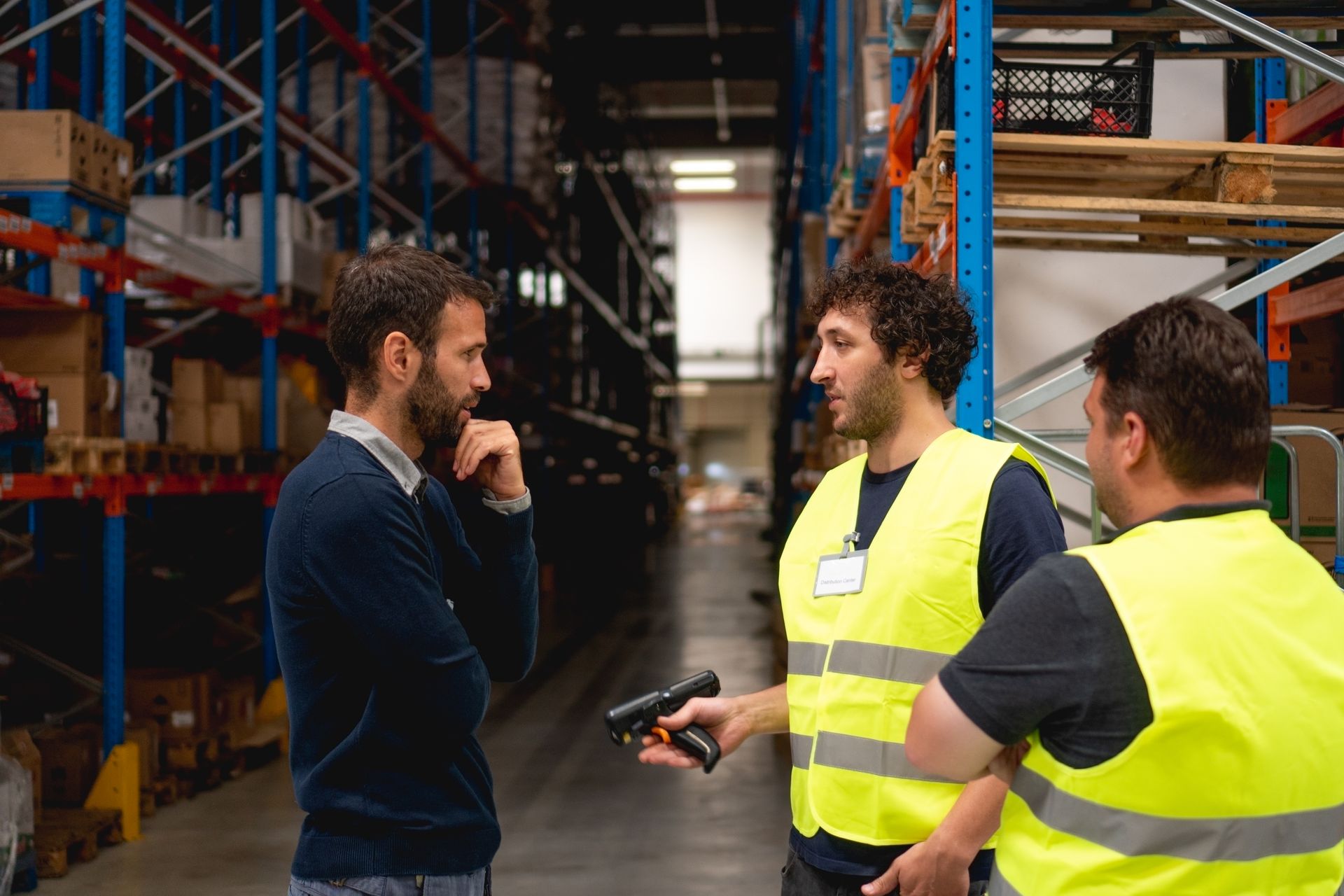 Three men in a warehouse; two wearing safety vests discuss with a man in a sweater.