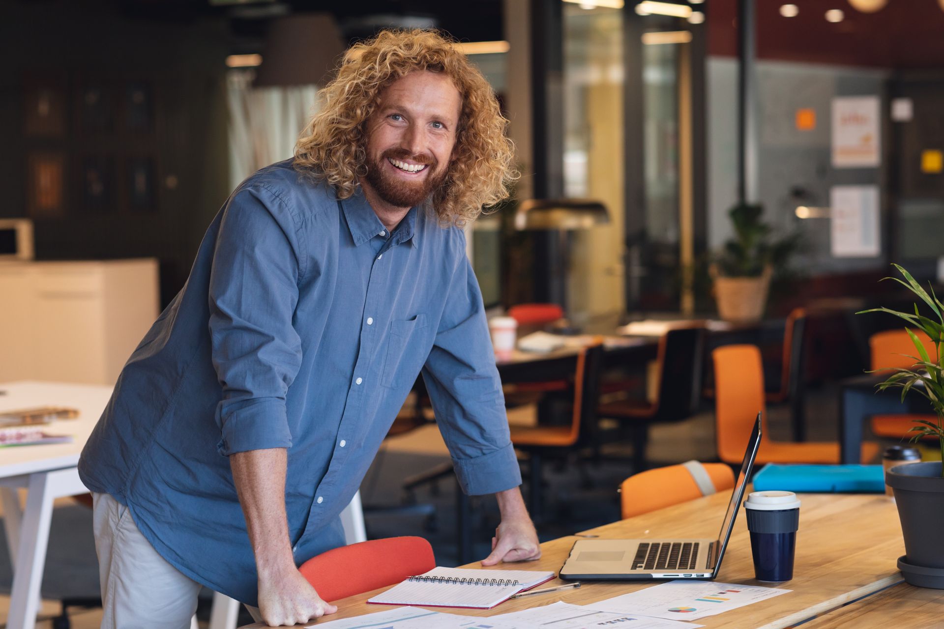 Man with curly hair smiles in an office, leaning on a desk with a laptop and papers.