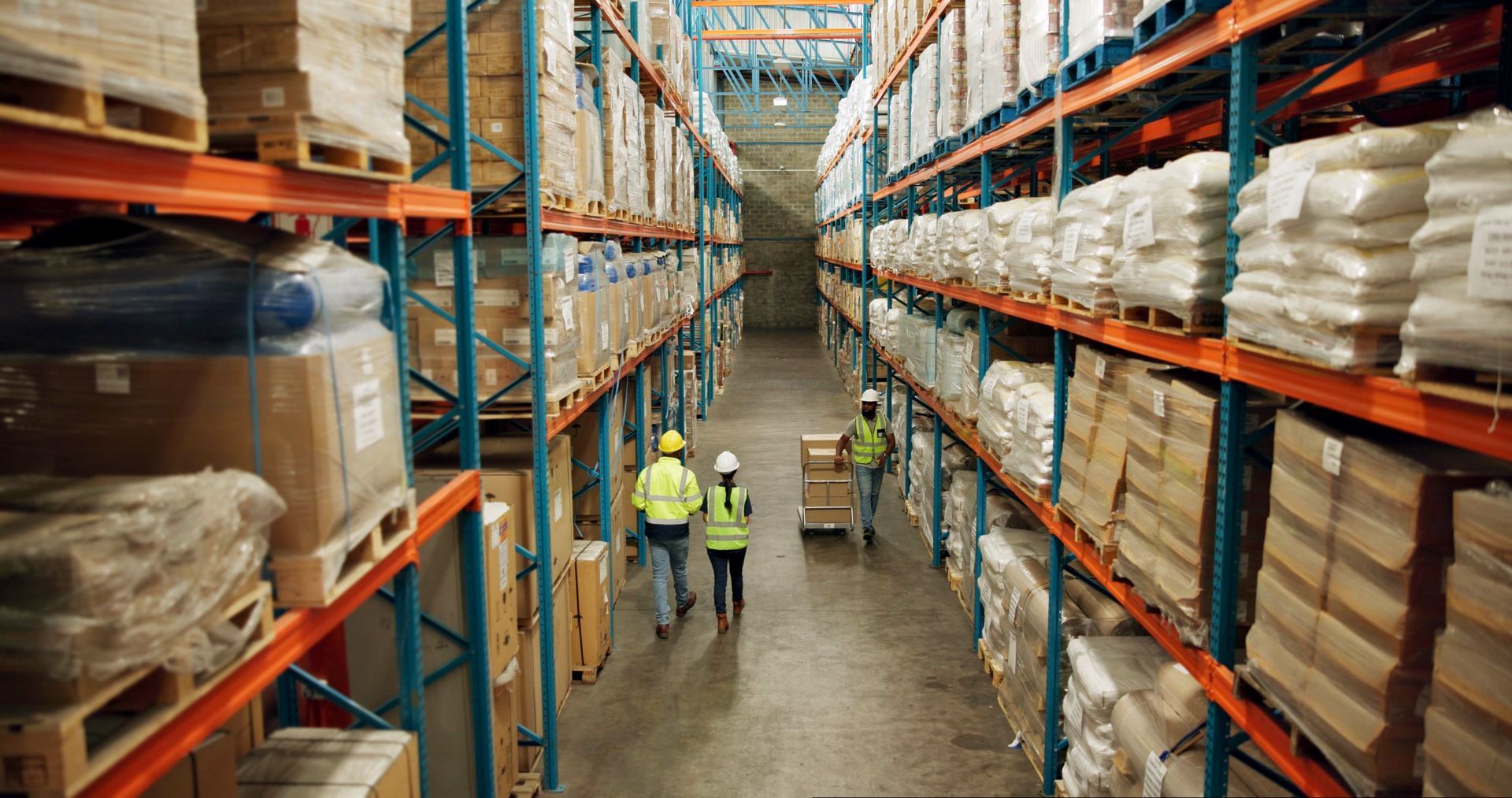 Warehouse interior with workers wearing safety vests, moving pallets and boxes on shelves.