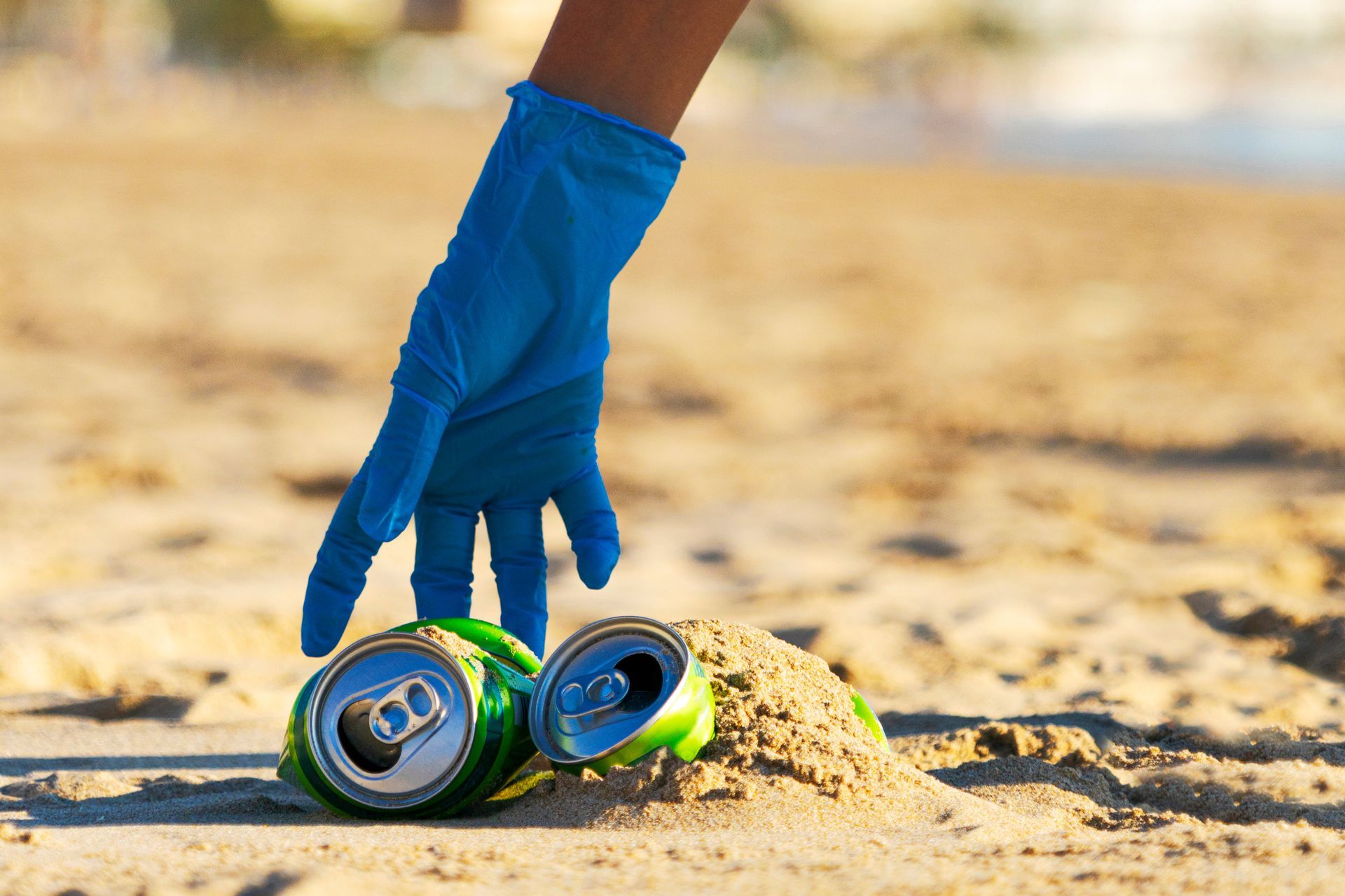 A close-up of a person in blue gloves collecting metal cans from a beach. A close-up of a person in blue gloves collecting metal cans from a beach.