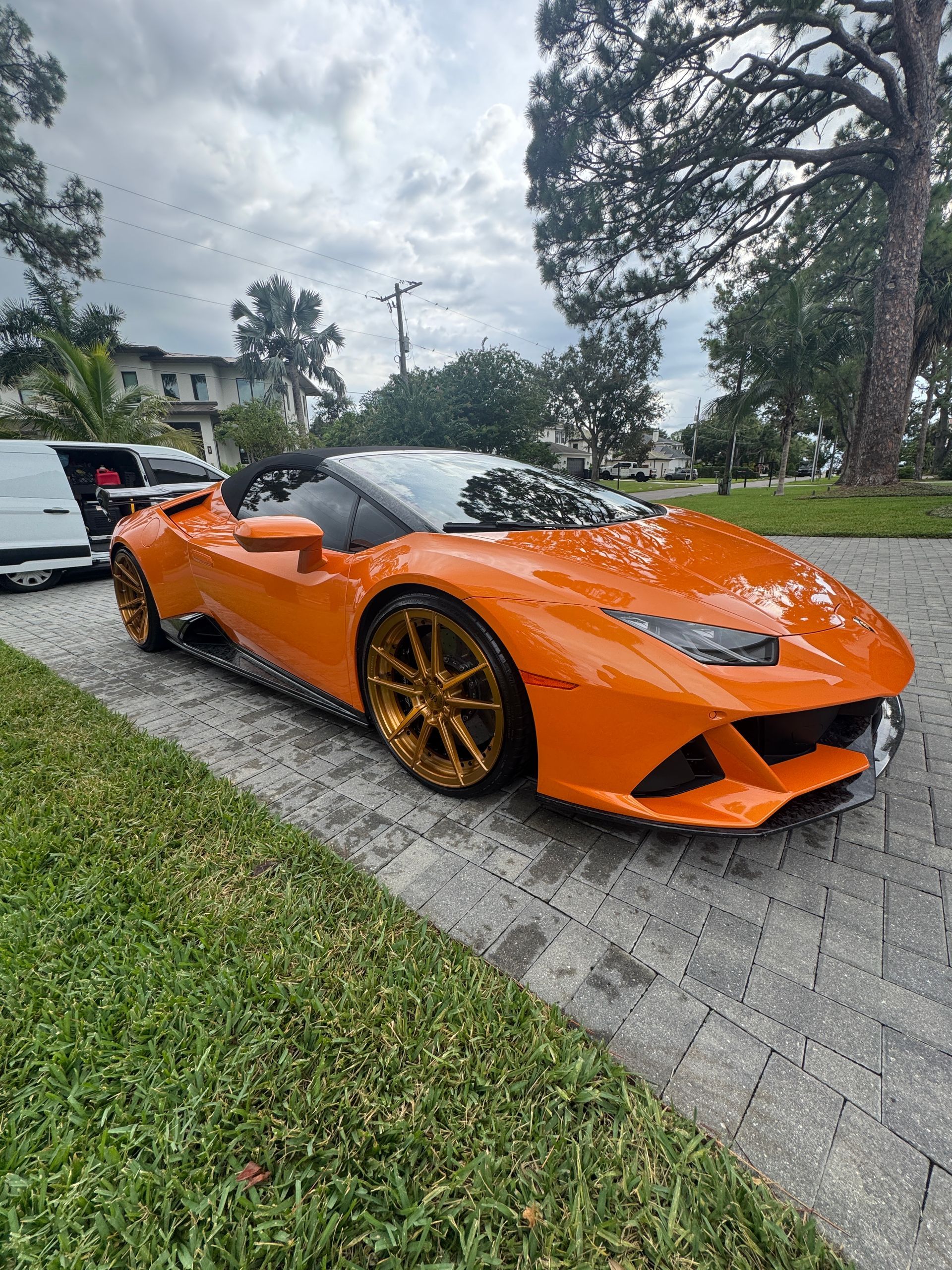 Orange Lamborghini sports car with gold rims parked on a brick driveway, next to green grass.