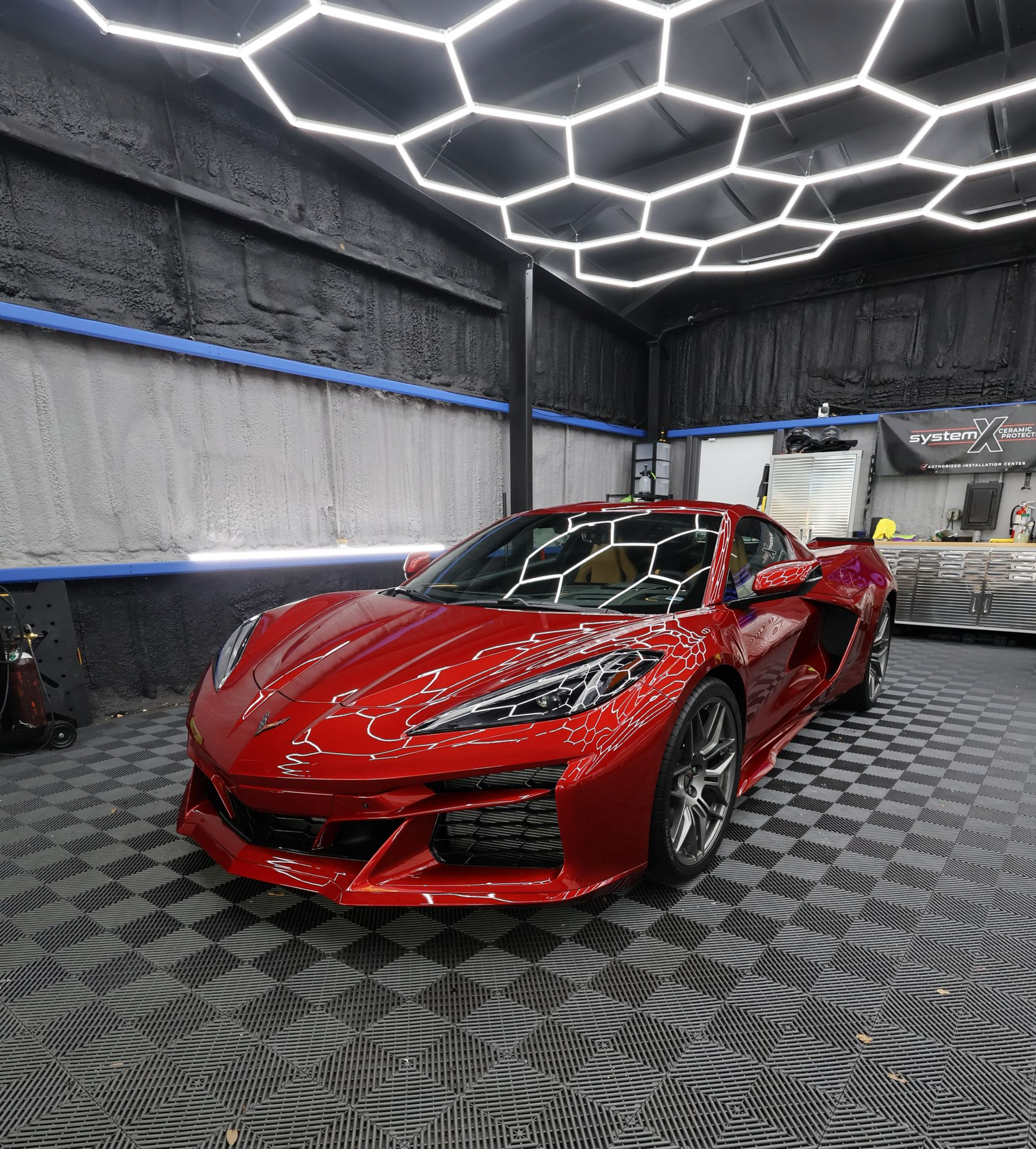 Red sports car in a garage with honeycomb ceiling lights, black and gray flooring.