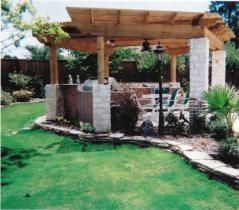 Outdoor kitchen under a wooden pergola with stone accents, nestled in a grassy backyard.