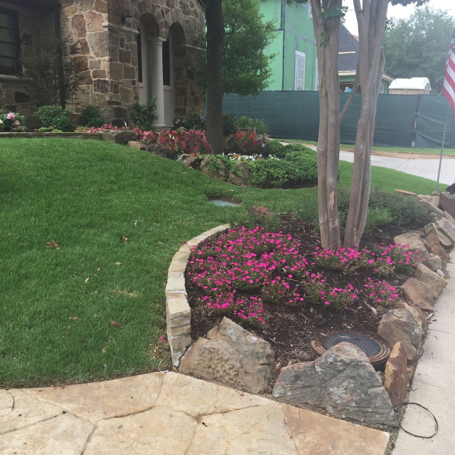 Stone-bordered flowerbeds filled with pink flowers and green shrubs in front of a house.