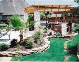 Backyard with pergola, fireplace, palm tree, landscaping, and green grass.