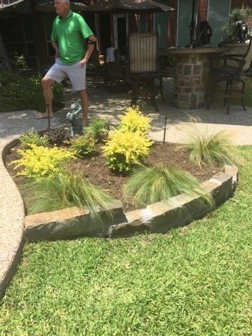 Man in green shirt stands near a curved flower bed with yellow and green plants, a stone border, and green grass.