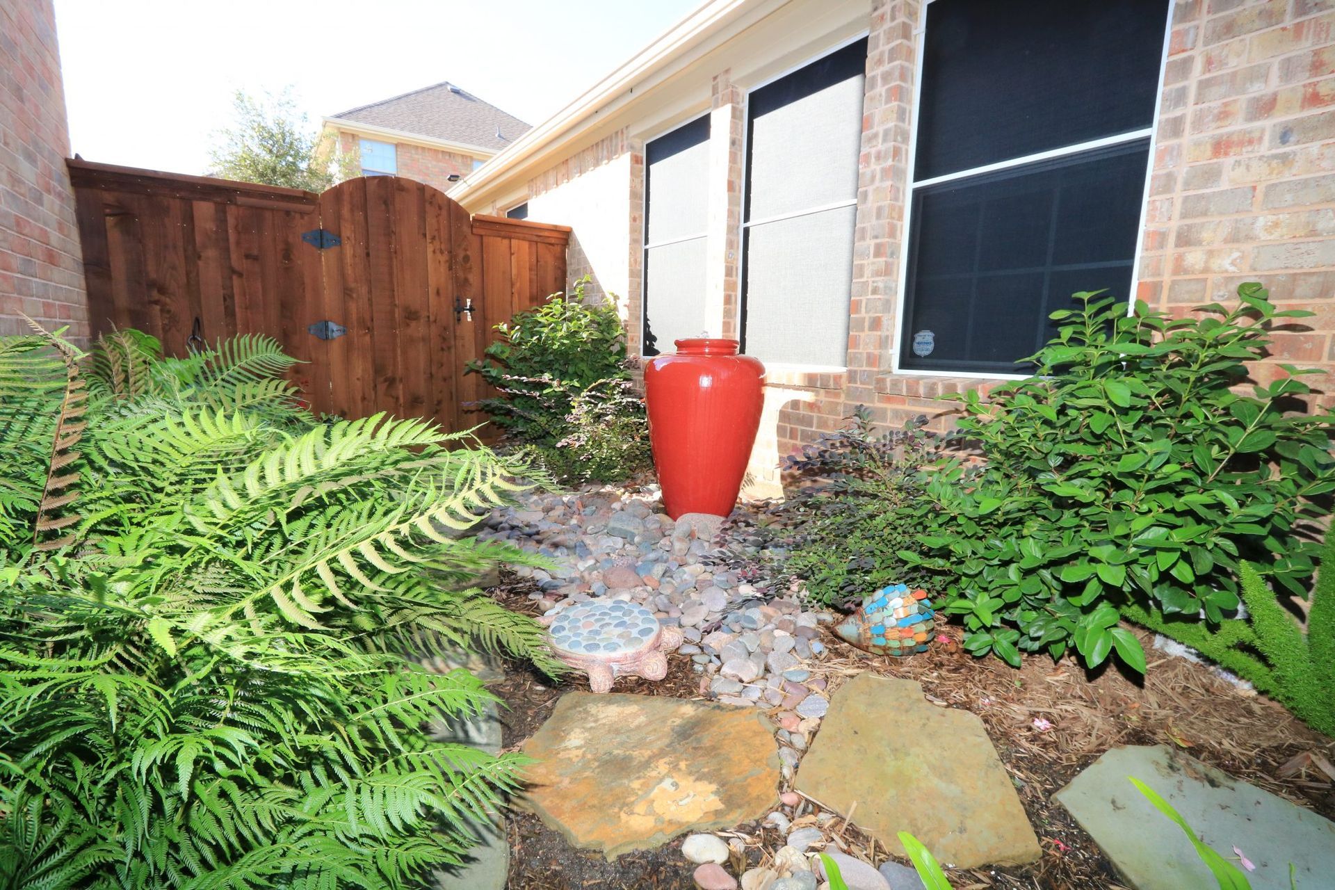 A small garden with a red vase, rocks, ferns, and shrubs, next to a brick house with a wooden gate.