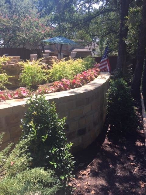 Stone retaining wall with flowers, greenery, and seating area with umbrella. American flag.