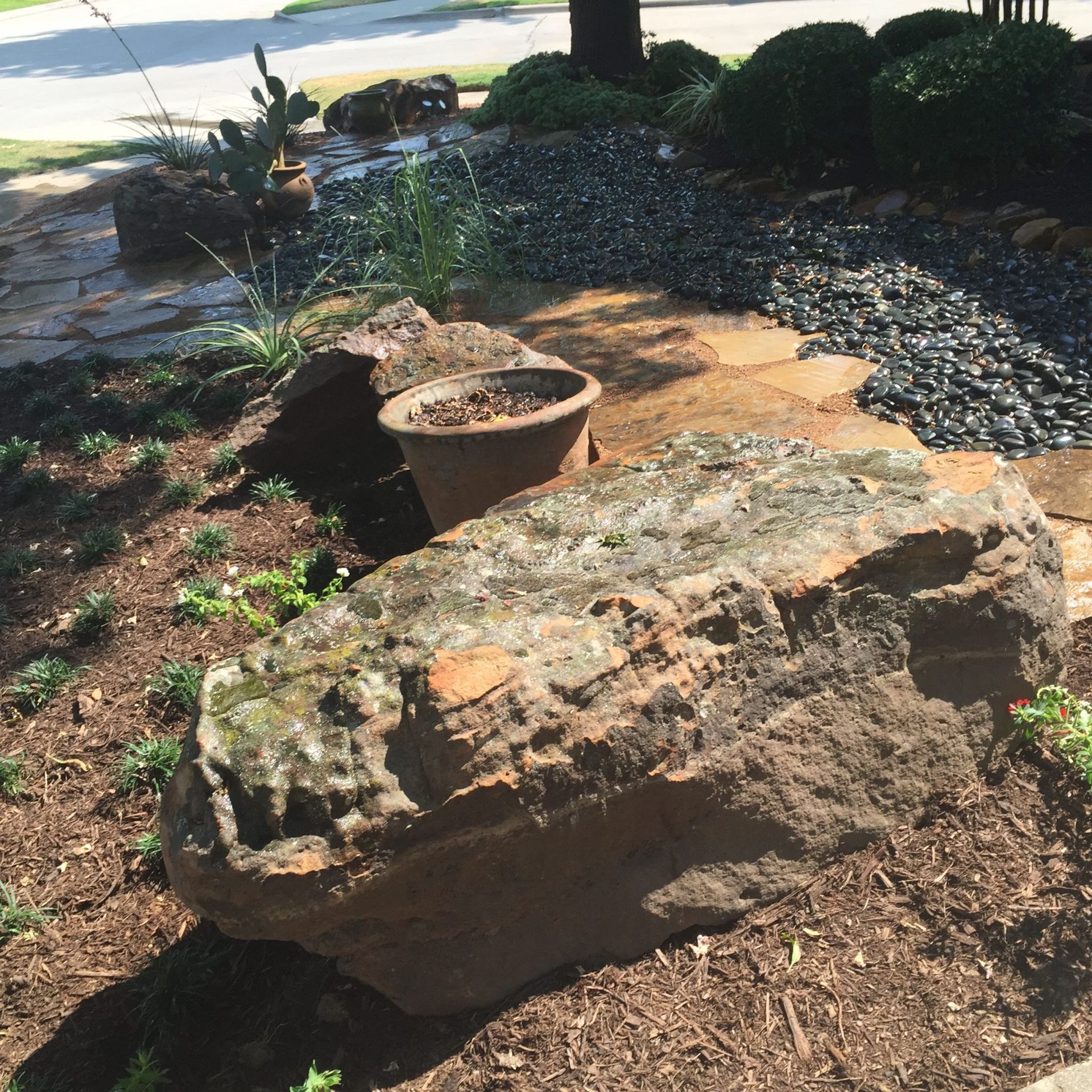Large weathered rock in a landscaped garden bed with mulch, small plants, and a terracotta pot.