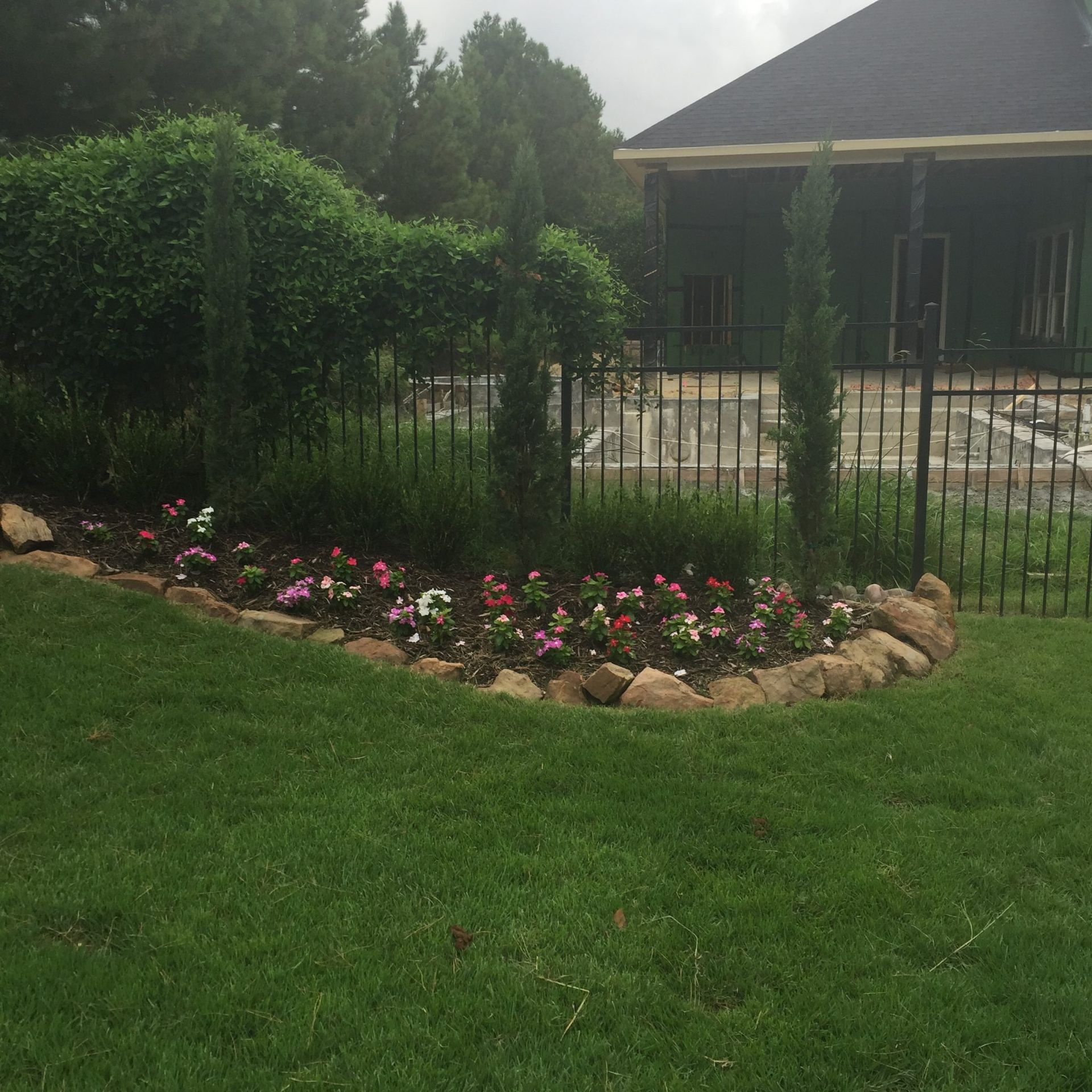 A colorful flower bed lined with rocks next to a black fence and a green lawn.