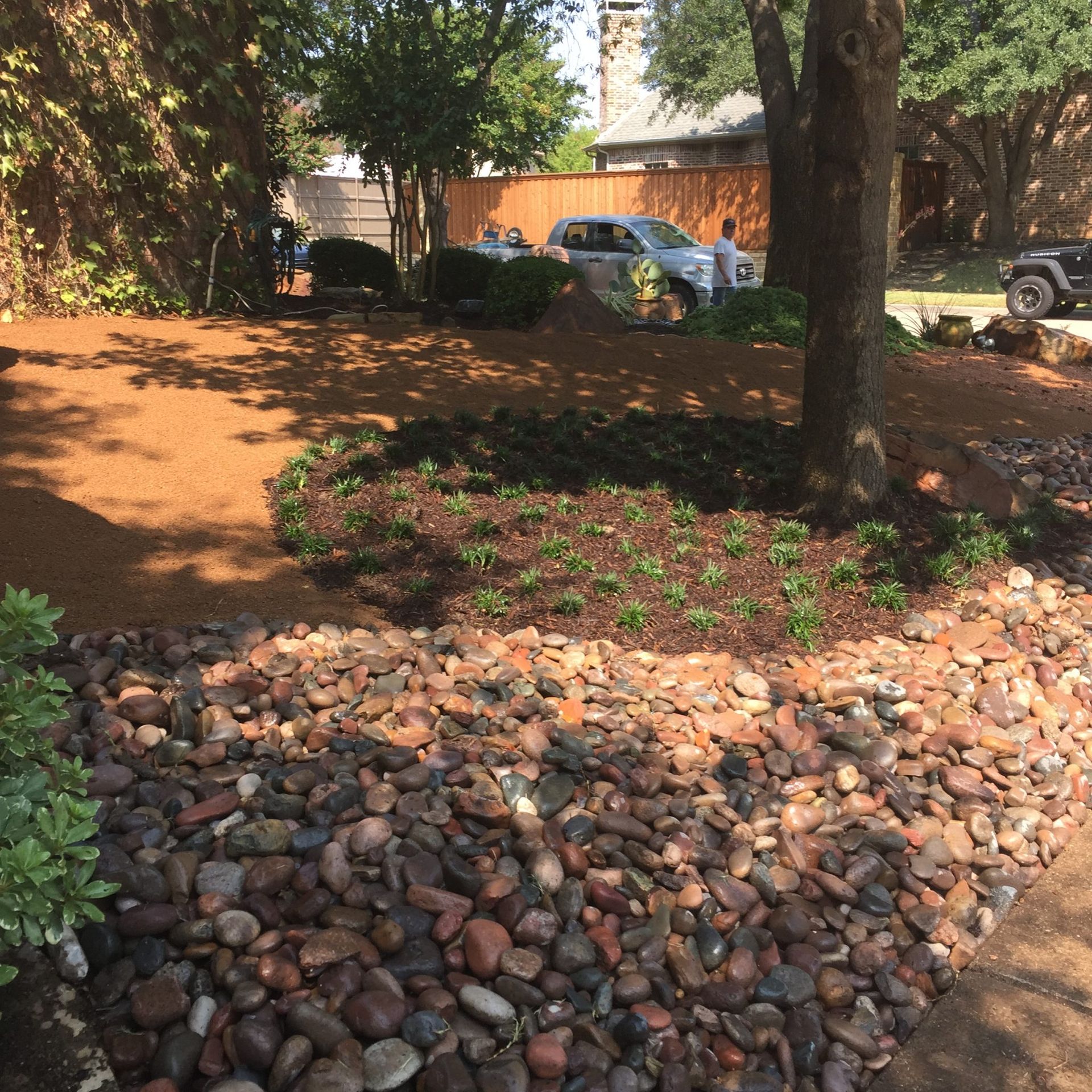 Landscape with mulch, plants, and rocks in a yard with a truck and trees.