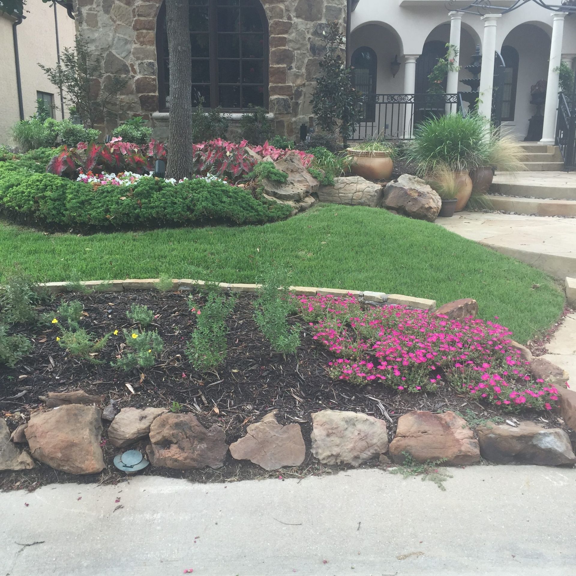 Landscaped front yard with stone beds, green grass, and colorful flowers in front of a stone house.