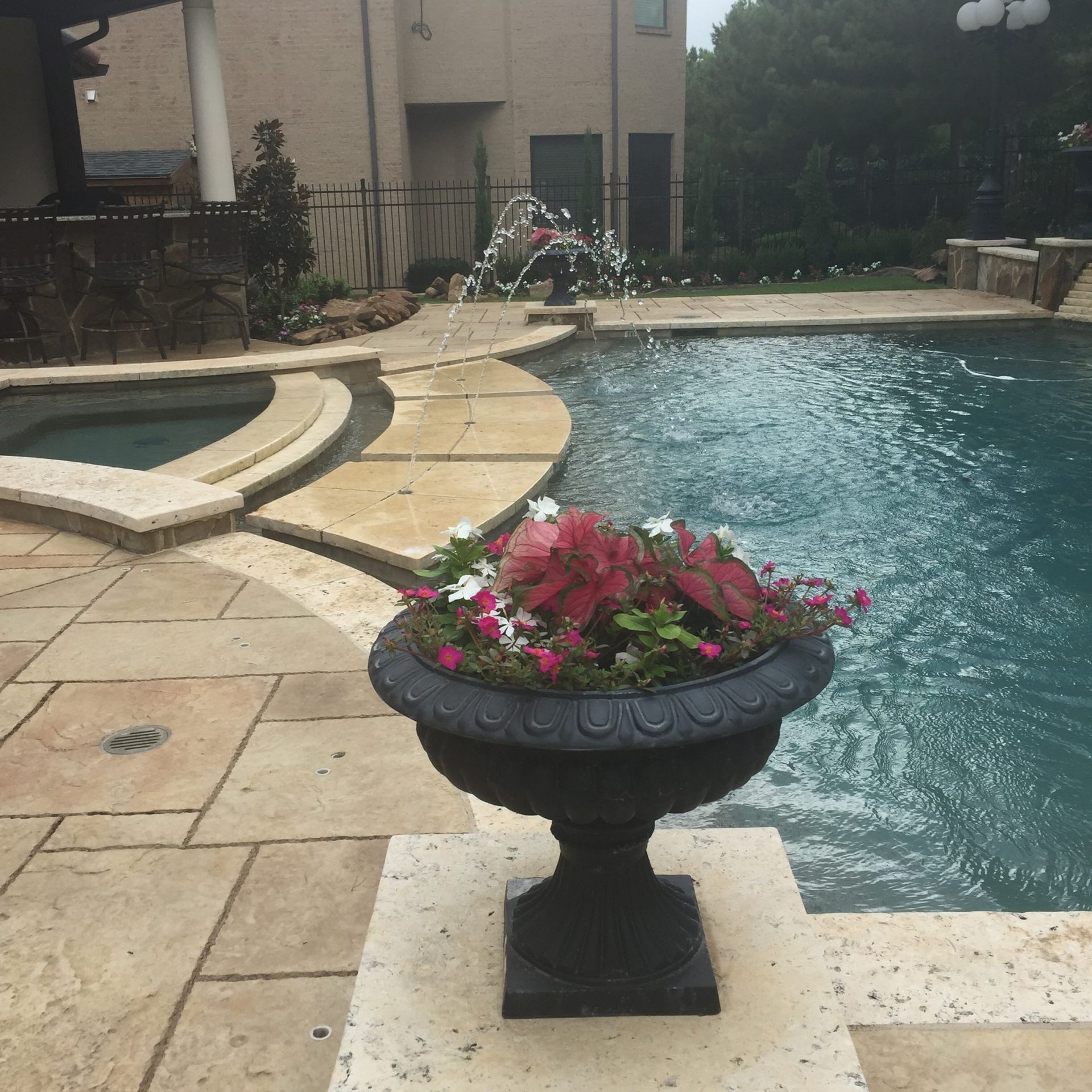 Black planter with flowers by a pool. Stone patio, fountain, and hot tub in the background.