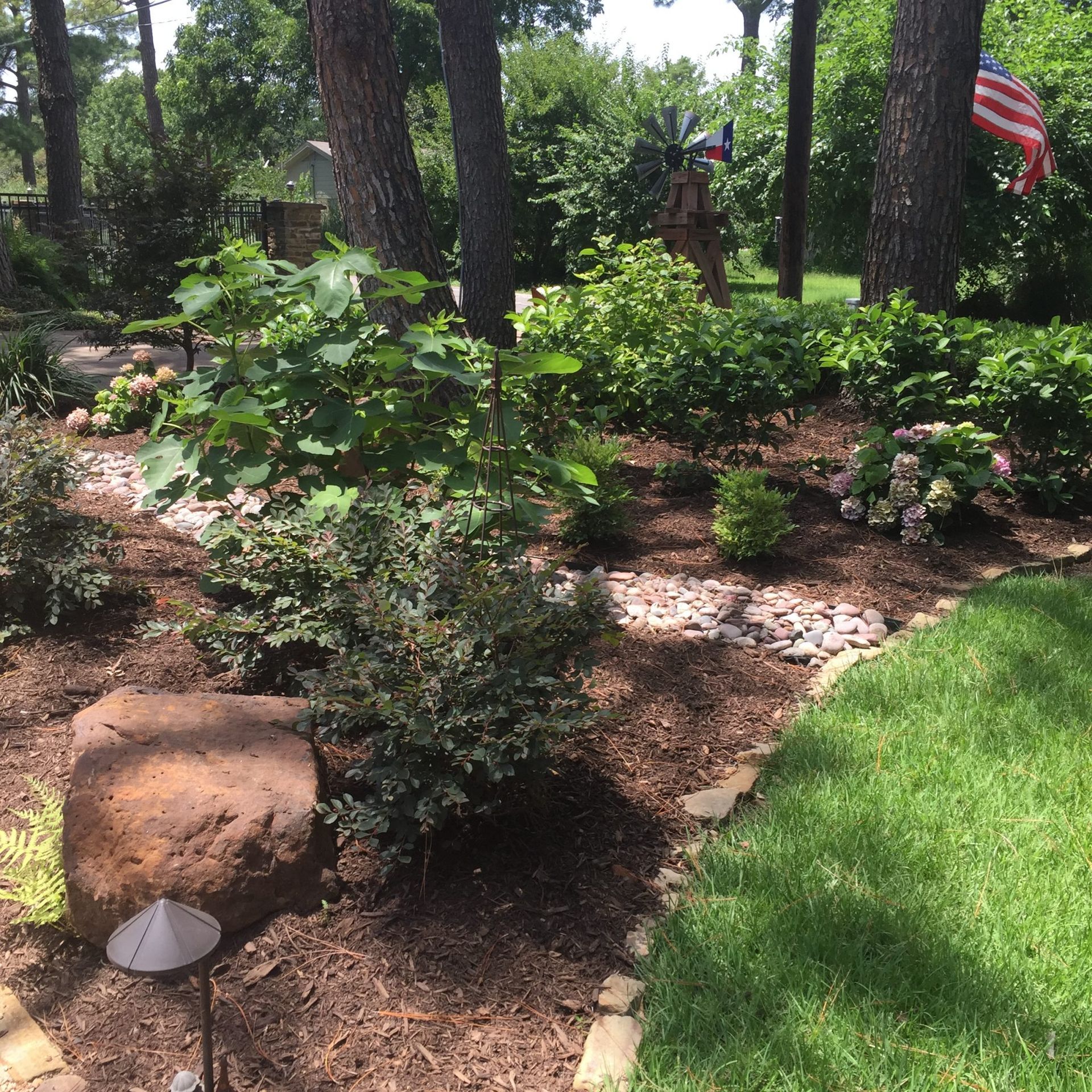 Lush garden with brown mulch, green plants, large rocks, and a neatly trimmed lawn with an American flag in the background.