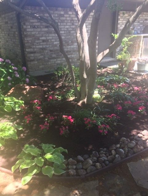 Garden bed with tree, pink flowers, hostas, rocks, and brick wall.