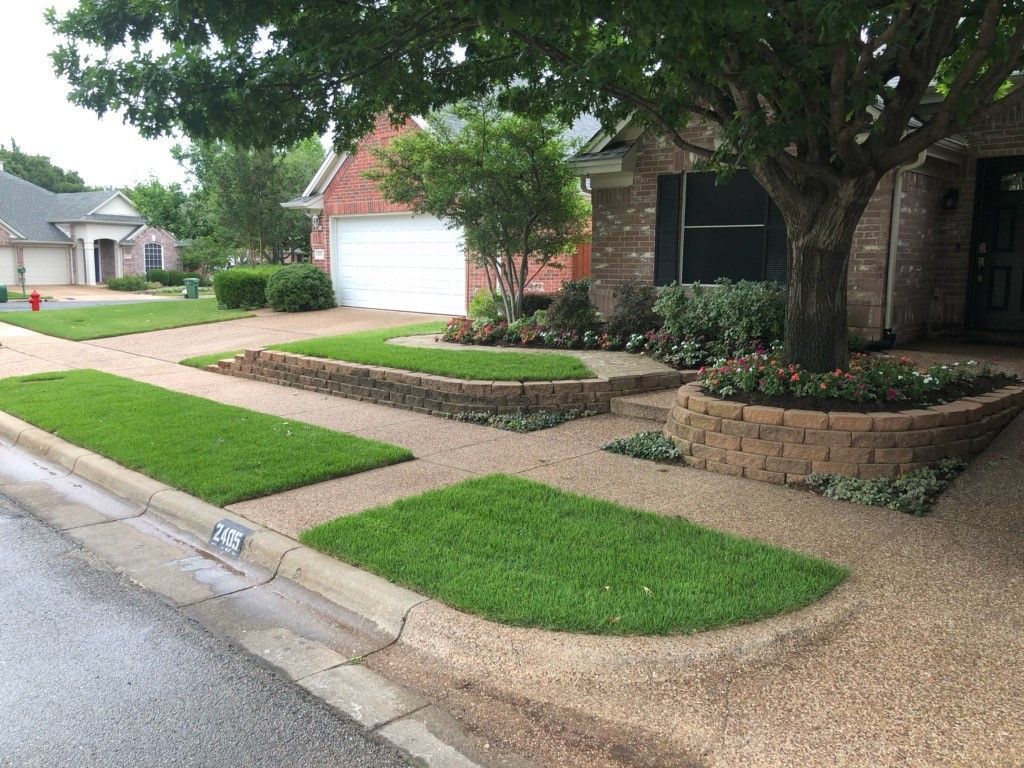 Residential yard with grass patches, gravel, and brick retaining walls. A tree is in front of the house.