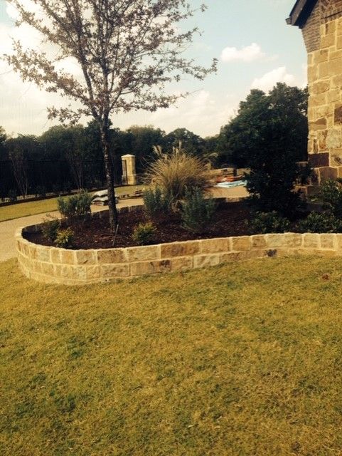 Stone-edged flower bed with mulch, plants, and a small tree in front of a house; green lawn.