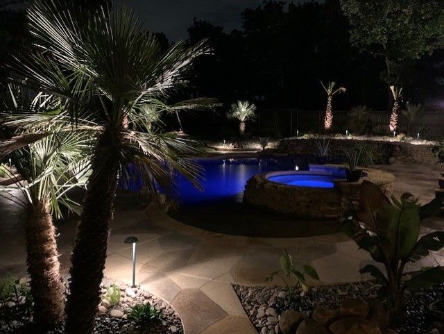 Nighttime view of a lit backyard with a pool, palm trees, and stone pathways; blue pool water reflects the lighting.