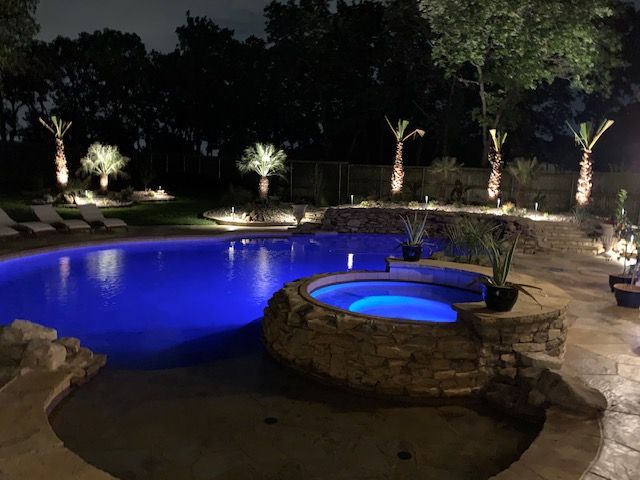 Night view of a pool and hot tub with blue and yellow lights, landscaped with palm trees.