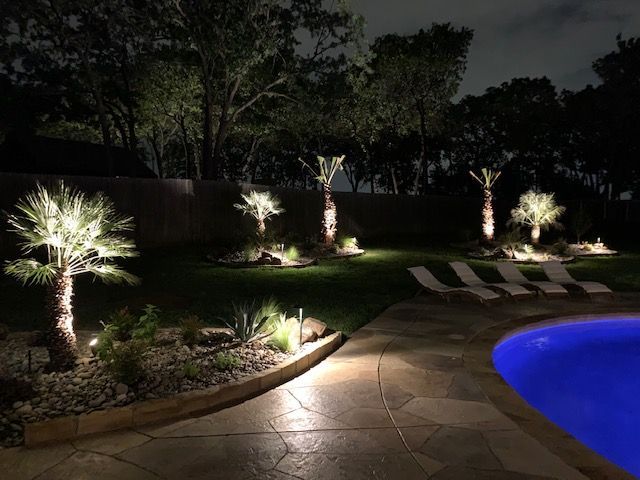 Nighttime backyard scene with illuminated palm trees, a pool, and lounge chairs.