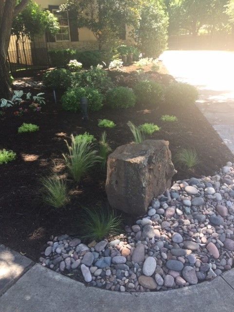 A rock garden with a large boulder, bordered by river stones and dark mulch with greenery.