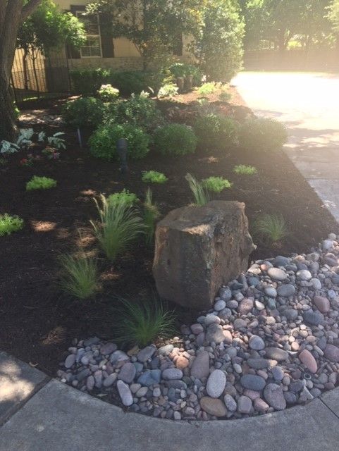 Landscaped yard with a large rock, plants, and river rocks bordering a sidewalk.
