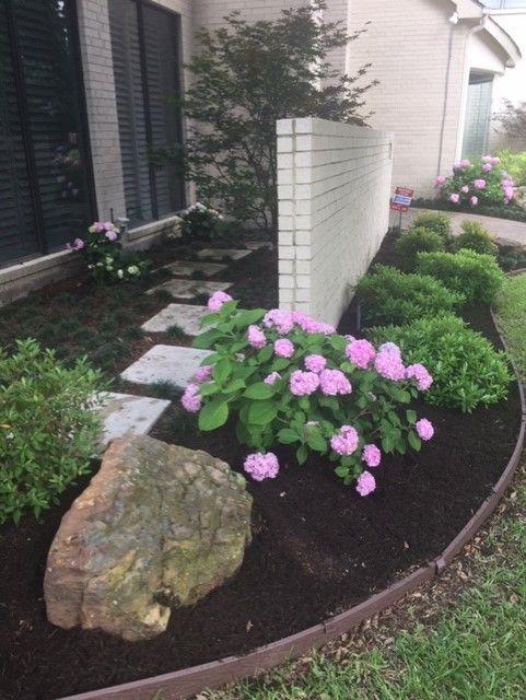 A landscaped yard with pink hydrangeas, stone pathway, and large rock in front of a brick wall.