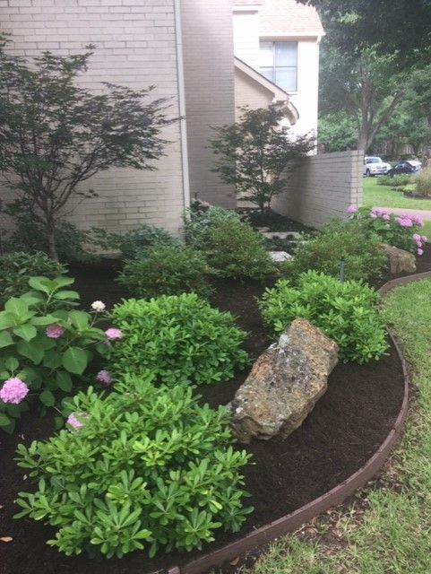 Landscaped flower bed with bushes, rocks, and mulch near a brick building.