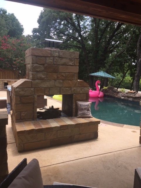 Stone fireplace next to a pool with pink flamingo float and umbrella, outdoor setting.