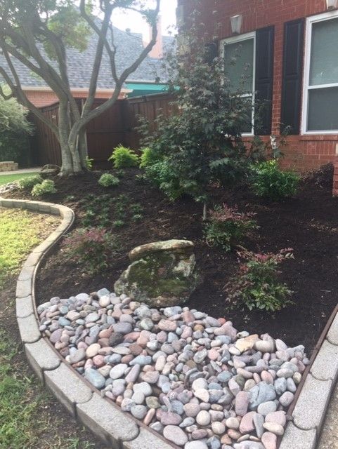 Landscaped front yard with a stone border, rock bed, and mulched flower bed with various plants.
