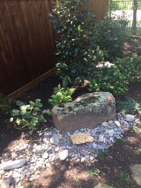 A stone fountain surrounded by rocks and plants in a garden setting next to a wooden fence.