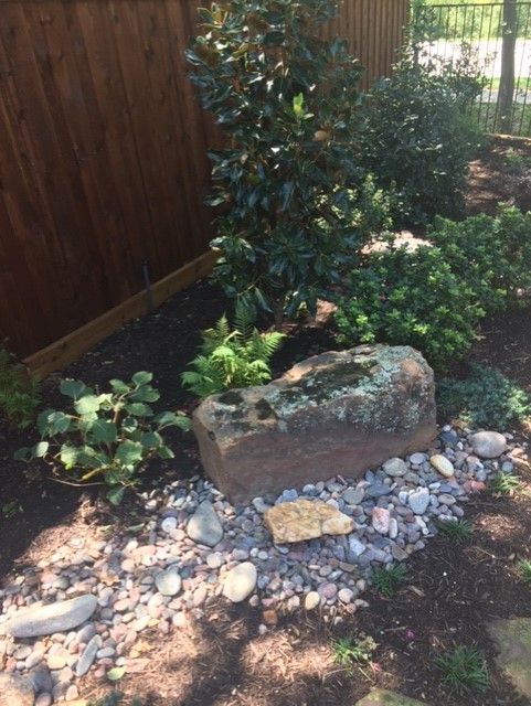 A weathered stone in a garden bed with various plants, next to a wooden fence.