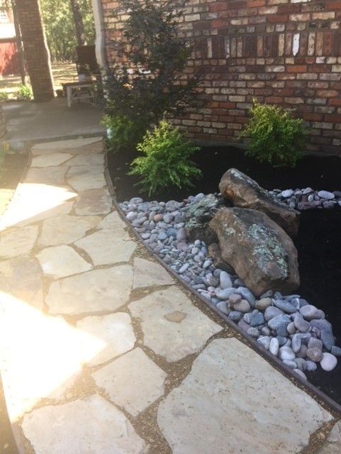 Flagstone walkway with a landscaped bed; rocks, mulch, and shrubs alongside a brick wall.